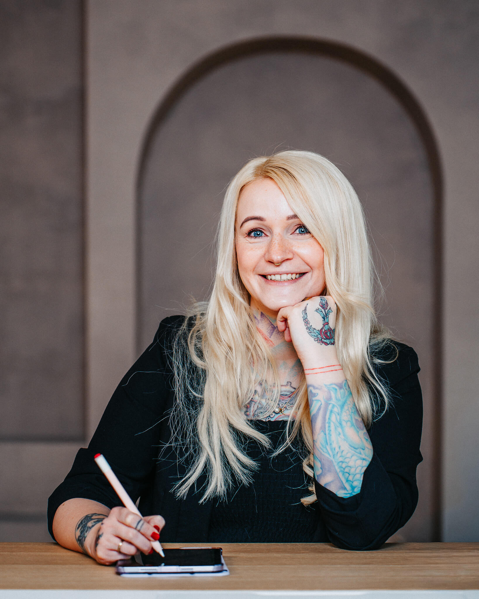 Porträtfoto von Christine Bach, Coachin, Speakerin und Beraterin für moderne Bildung, digitale Transformation und nachhaltigen Wandel. Sie sitzt lächelnd an einem Tisch, hält einen Stift in der Hand und trägt ein schwarzes Oberteil. Ihre Unterarme sind mit Tattoos verziert.