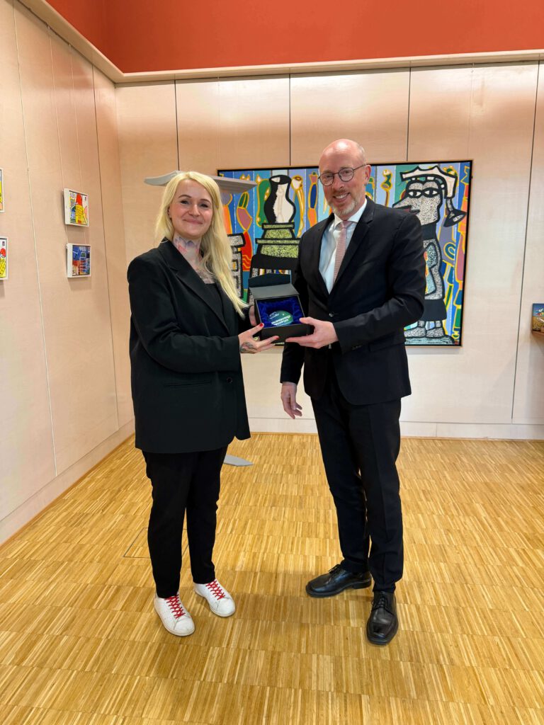 Christine Bach und Christian Pegel, Minister für Inneres und Bau aus Mecklenburg-Vorpommern, halten gemeinsam eine schwarze Box mit blauem Licht in einem Raum mit Holzfußboden und bunter Wandkunst. Christine Bach wurde zur Digitalisierungsbotschafterin MV ausgezeichnet.