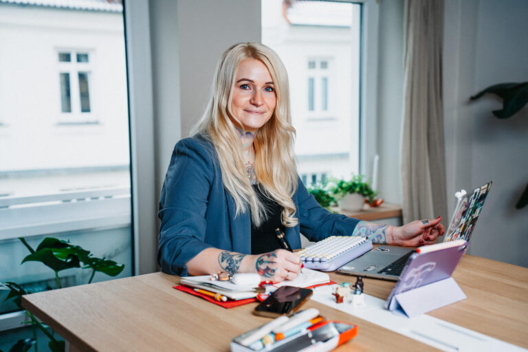 Christine Bach, Coachin, Speakerin und Beraterin für moderne Bildung, digitale Transformation und nachhaltigen Wandel, sitzt konzentriert an einem Tisch. Sie schreibt Notizen in ein Buch und trägt ein dunkles Blazer-Oberteil. Auf dem Tisch stehen ein Laptop und ein Tablet, beide mit zahlreichen Aufklebern verziert. Ihre Arme sind mit Tattoos geschmückt.