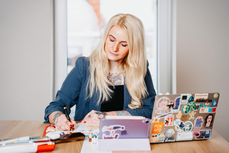 Christine Bach, Coachin, Speakerin und Beraterin für moderne Bildung, digitale Transformation und nachhaltigen Wandel, sitzt konzentriert an einem Tisch. Sie schreibt Notizen in ein Buch und trägt ein dunkles Blazer-Oberteil. Auf dem Tisch stehen ein Laptop und ein Tablet, beide mit zahlreichen Aufklebern verziert. Ihre Arme sind mit Tattoos geschmückt.
