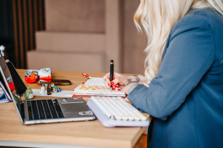 Eine Person mit langen blonden Haaren sitzt an einem Tisch und schreibt konzentriert in ein Notizbuch. Auf dem Tisch stehen ein Laptop mit einem Tablet-Ständer, ein Taschenrechner und verschiedene Stifte. Die Person trägt Tattoos auf den Armen.