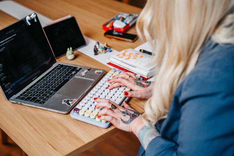 Christine Bach, Coachin, Speakerin und Beraterin für moderne Bildung, digitale Transformation und nachhaltigen Wandel, sitzt an einem Schreibtisch und arbeitet an einer mechanischen Tastatur. Vor ihr steht ein Laptop mit geöffnetem Code-Editor, und auf dem Tisch liegen verschiedene Arbeitsutensilien wie ein Notizbuch, Stifte und kleine Figuren. Ihre Arme sind mit Tattoos verziert.