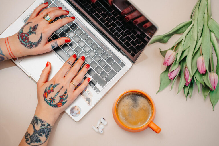 Eine Person mit Tattoos auf den Armen tippt auf der Tastatur eines Laptops. Auf dem Tisch liegen neben dem Laptop ein orangefarbener Kaffeebecher, weiße In-Ear-Kopfhörer und ein Strauß mit rosa und grünen Tulpen.